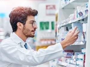 Farmacêutico de jaleco e óculos lendo atentamente as informações de uma caixa de medicamento em uma prateleira de farmácia, ilustrando a importância de verificar o registro Anvisa.