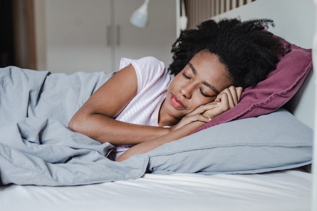 Mulher negra deitada de lado, dormindo de forma serena e profunda em uma cama confortável com lençóis cinza, representando o objetivo final de praticar a higiene do sono.