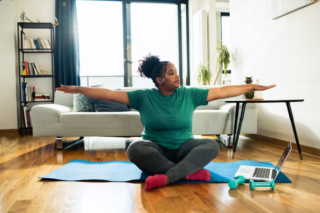 Mulher negra sentada com as pernas cruzadas em um tapete de yoga azul na sala de estar, com os braços abertos, preparando-se para praticar exercícios para fazer em casa enquanto acompanha uma aula pelo notebook.