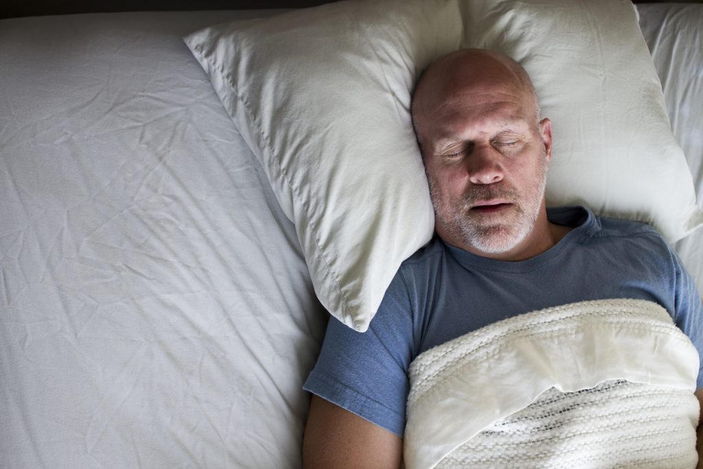 Homem calvo dormindo em uma cama com travesseiros brancos e roupas de cama claras, ilustrando o tema da apneia do sono e a busca pela qualidade do descanso.