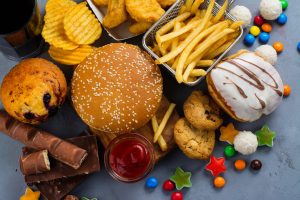Fotografia vista de cima de uma grande quantidade de alimentos ultraprocessados misturados. Há um hambúrguer com gergelim, porções de batata frita, salgadinhos, nuggets, barras de chocolate, um donut com cobertura, biscoitos e balas, representando os tipos de alimentos frequentemente consumidos em um episódio de compulsão alimentar.