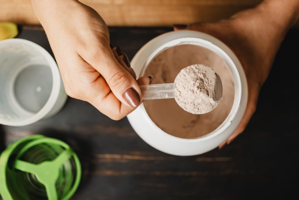 Visão superior de mãos segurando um medidor (scoop) cheio de suplemento em pó sobre um pote branco. A imagem ilustra o guia principal que explica para que serve whey e como inseri-lo de forma prática no dia a dia.